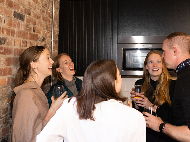 A group of five people stand in a kitchen, engaging in lively conversation. They are holding drinks and smiling, with brick walls and kitchen appliances visible in the background. They appear to be enjoying each other's company.