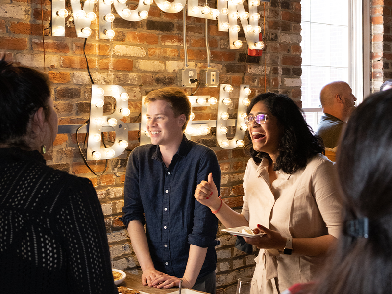 A group of people enjoy a conversation in a warmly lit brick-walled space. Two individuals, one in a blue shirt and the other in a light-colored outfit, are laughing and engaged with others. Illuminated signage in the background spells "MOON BREW.