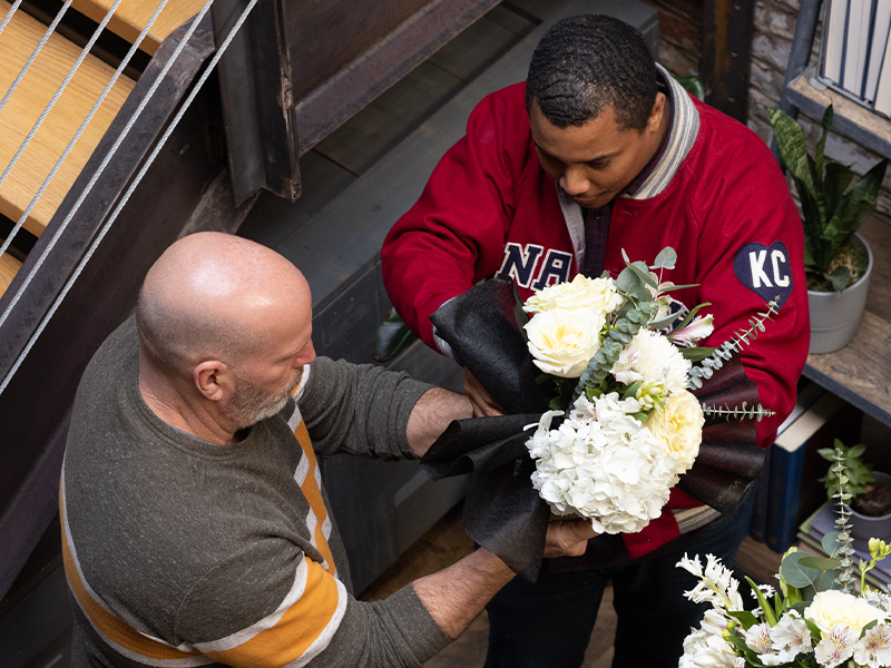 Two men, one with a bald head and wearing a striped sweater, and the other with short hair, a red jacket, and "KC" on the sleeve, collaborate on arranging a bouquet of white flowers and greenery. They are indoors, near a staircase and bookshelf.