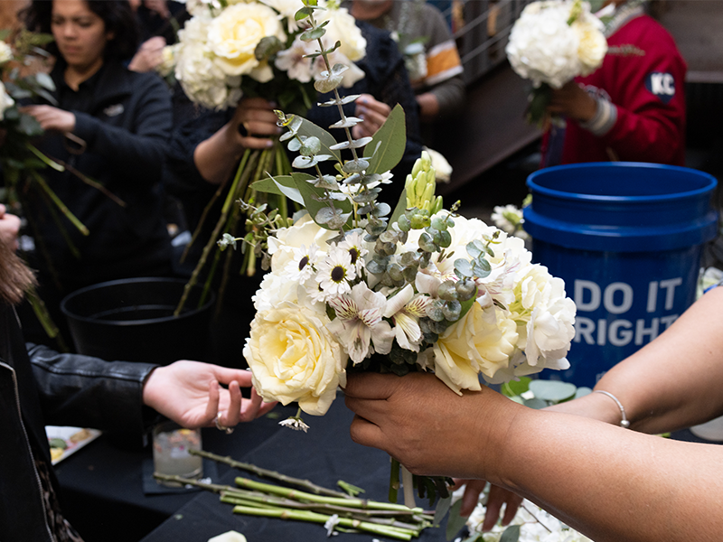 People arranging white flower bouquets at a table. The scene is lively with multiple hands holding and arranging flowers. A blue bucket with the words "DO IT RIGHT" and various flower stems are visible on the table in the background.