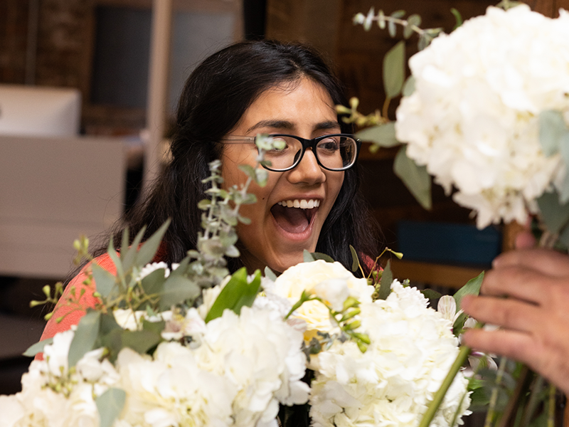 A woman with long dark hair and glasses smiles joyfully while arranging a bouquet of white flowers. She is indoors, and the background is slightly blurred, focusing attention on her happy expression and the flowers.