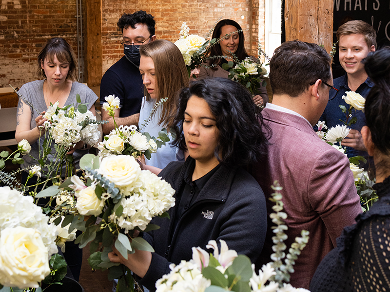 A group of people gather in a bright room, arranging bouquets of white flowers. Some wear casual attire, while one person in a pink blazer stands out. The atmosphere appears focused and collaborative, with a brick wall in the background.