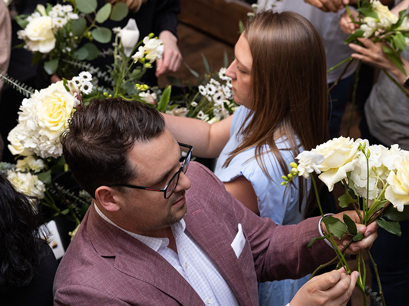 A group of people are arranging floral bouquets. A man in a pink blazer holds white flowers and greenery, concentrating on his work. A woman beside him, in a blue shirt, arranges her bouquet. Other individuals around them are similarly focused on their flowers.