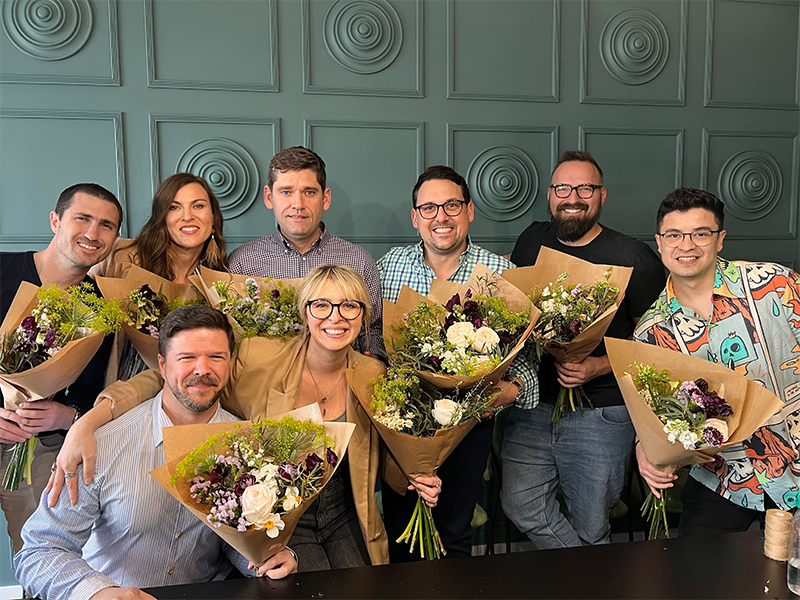 A group of eight people standing close together, smiling, and holding bouquets of flowers. The background features a green, decorative wall. The individuals appear to be enjoying a celebratory moment.
