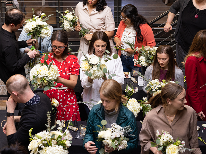 A group of people gather around tables, each arranging white and cream-colored flowers into bouquets. They are focused on their tasks, and the setting appears to be indoors with a mix of casual and business casual attire.