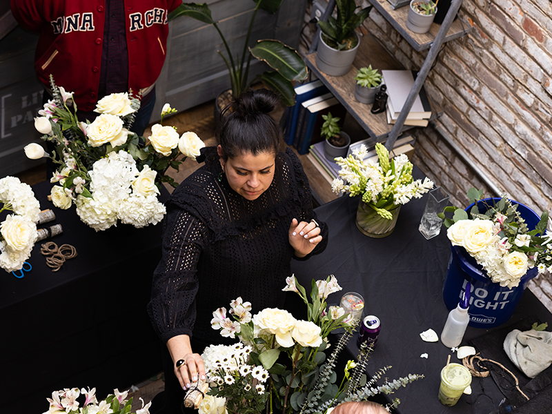 A woman in a black lace top arranges a floral bouquet at a table filled with various flowers. She is surrounded by shelves with plants and books. Another person in a red jacket is partially visible in the background. The scene is set in a rustic, brick-walled room.