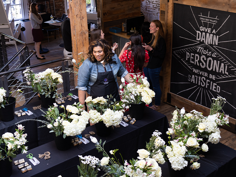 A florist's workshop with several people. A woman stands behind a table filled with white flower arrangements. Other attendees, including someone in a red dress, engage in conversation nearby. A blackboard on the wall displays motivational quotes in white chalk.