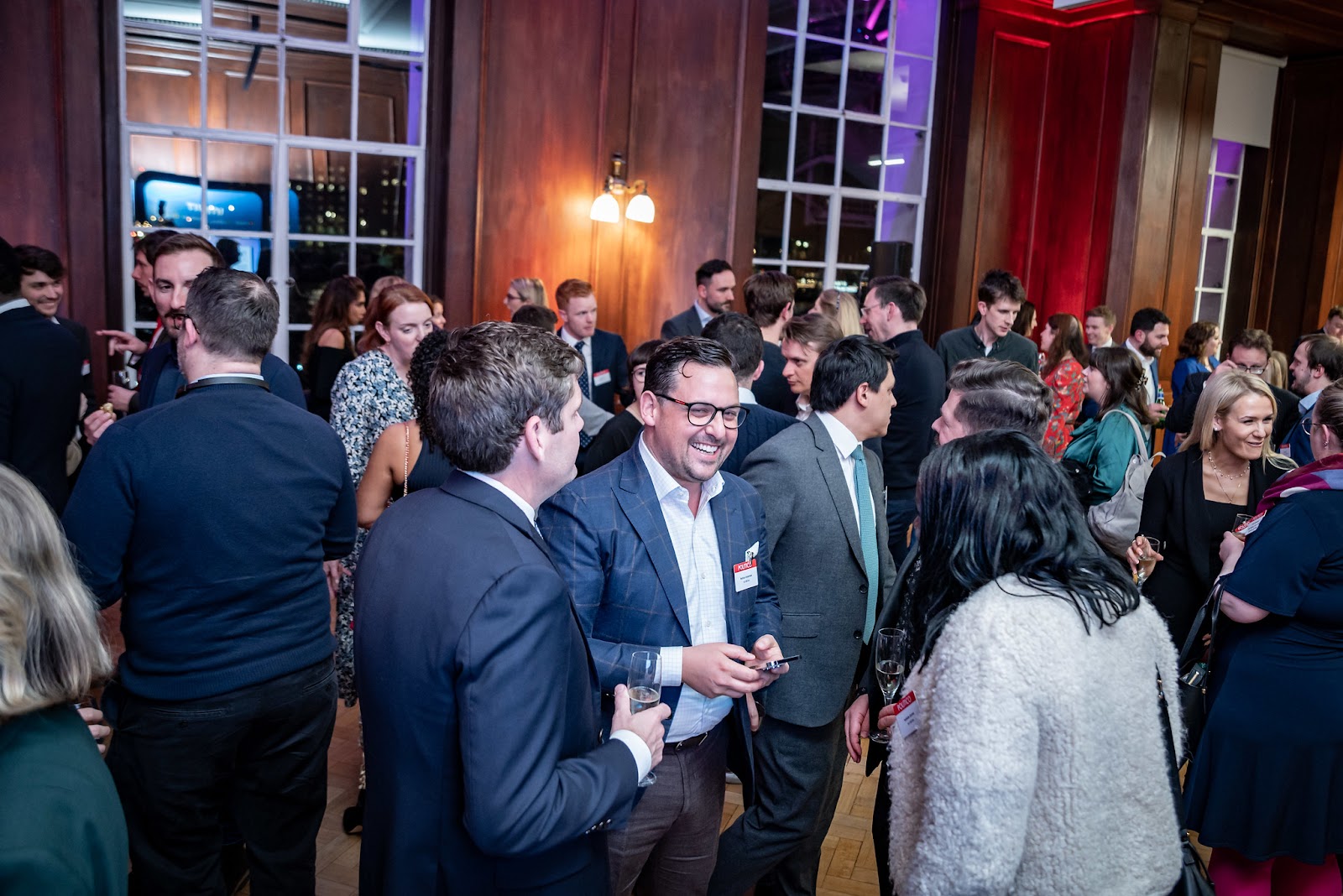 A large group of people socializing at an indoor event. Men and women are engaged in conversations, holding drinks, and some are smiling or laughing. The room has wooden paneled walls and large windows, and is lit with a combination of warm and colored lighting.