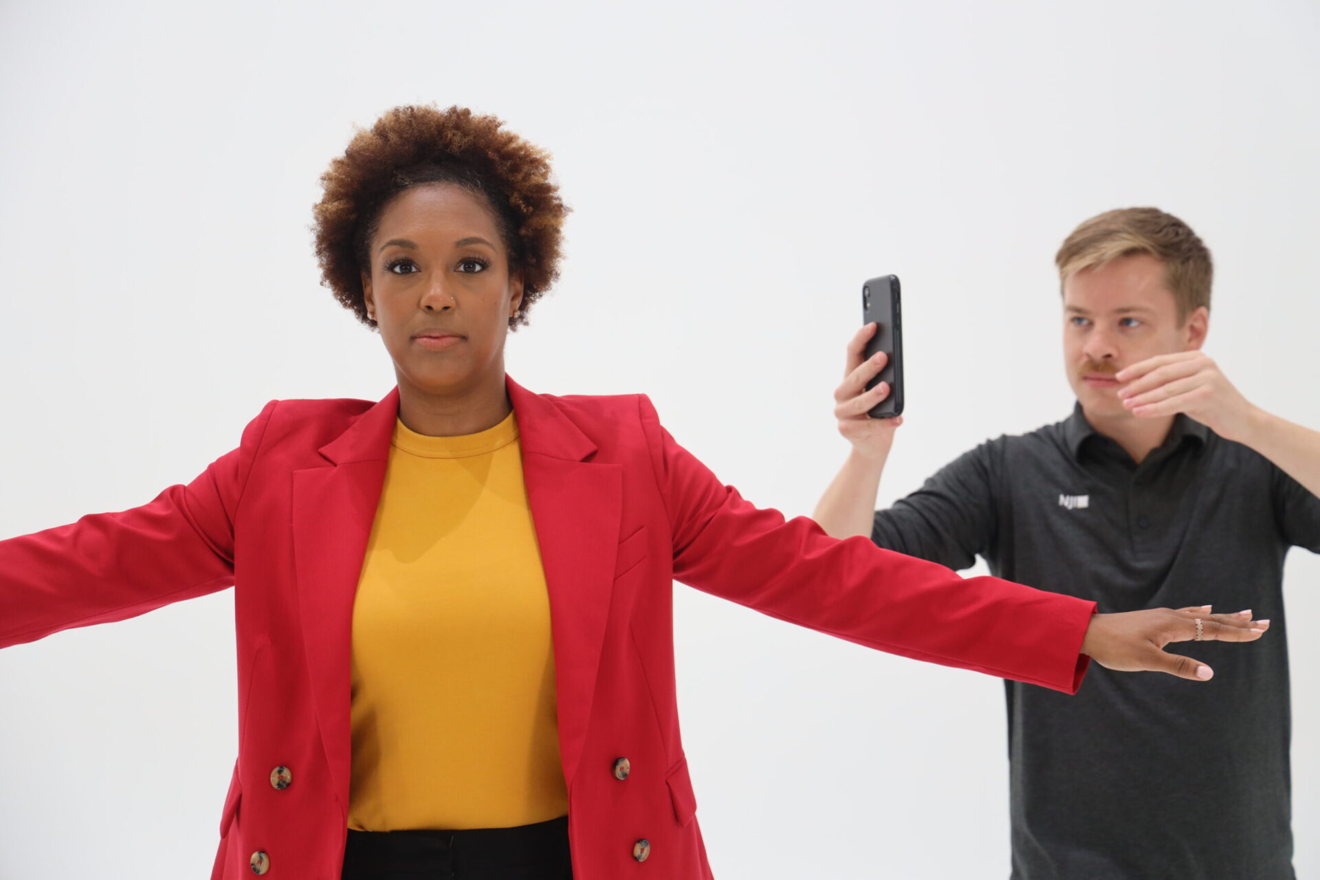 A woman wearing a red blazer and yellow top stands with her arms outstretched against a white background. A man beside her uses a smartphone, holding it up as if to photograph or scan her.