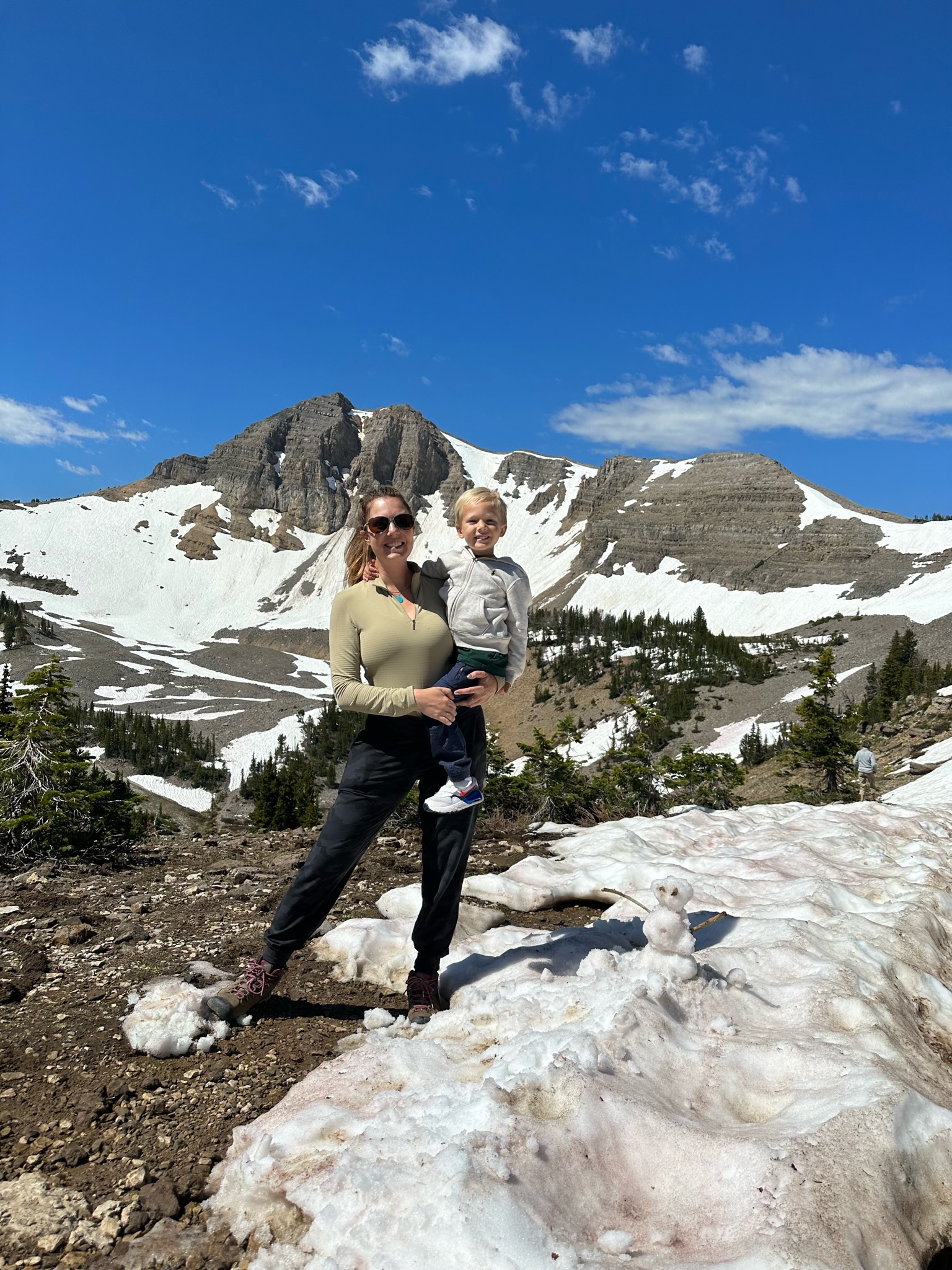 A woman holding a young child stands on a snow-covered mountain with patches of grass and conifer trees around. Rocky peaks are prominent in the background, set against a bright blue sky with scattered clouds.