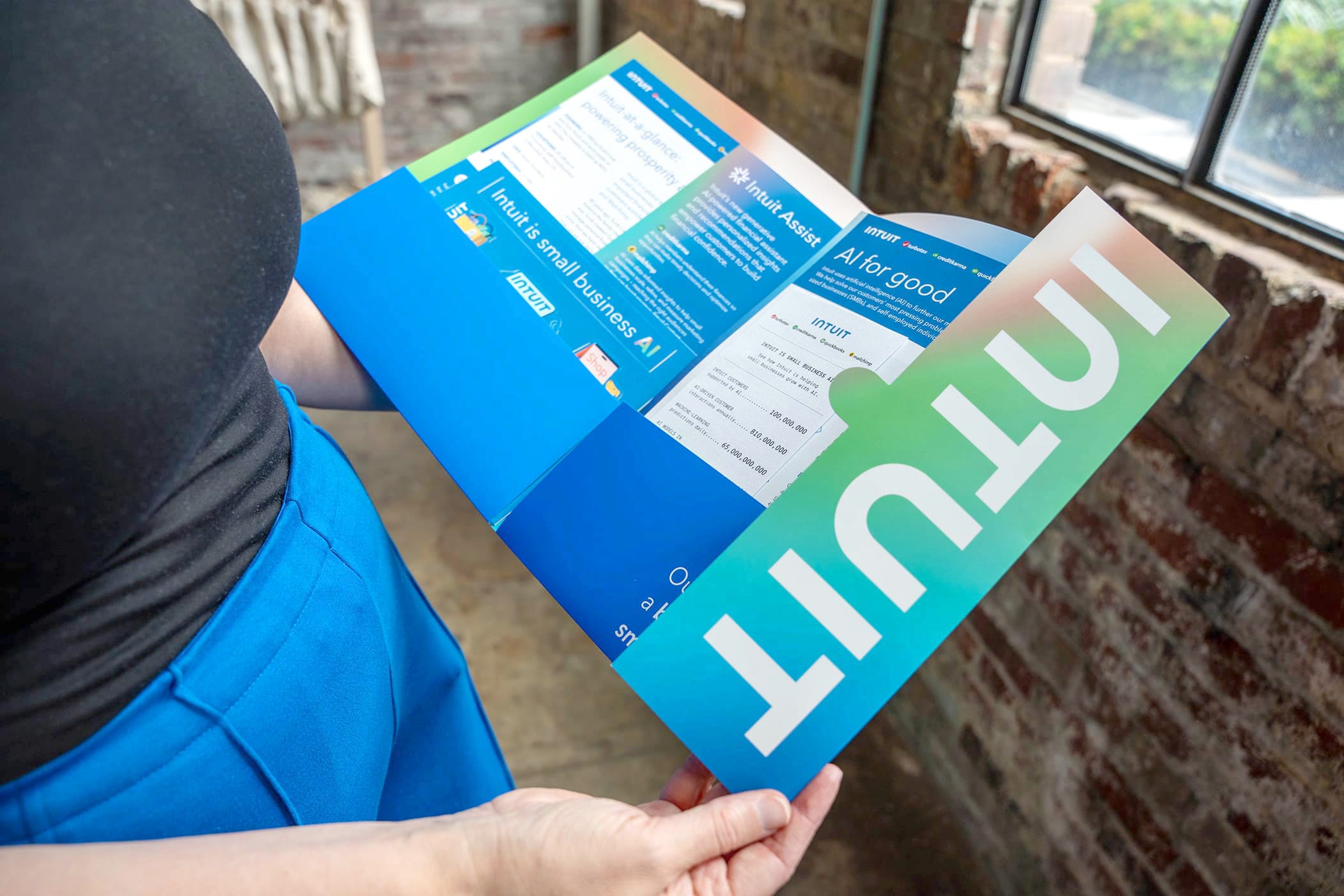 A person in a black shirt and blue pants is holding and opening a colorful pamphlet with the word "INTUIT" prominently displayed. The pamphlet has text and images, indicating it contains informational content. They are near a brick wall with windows.