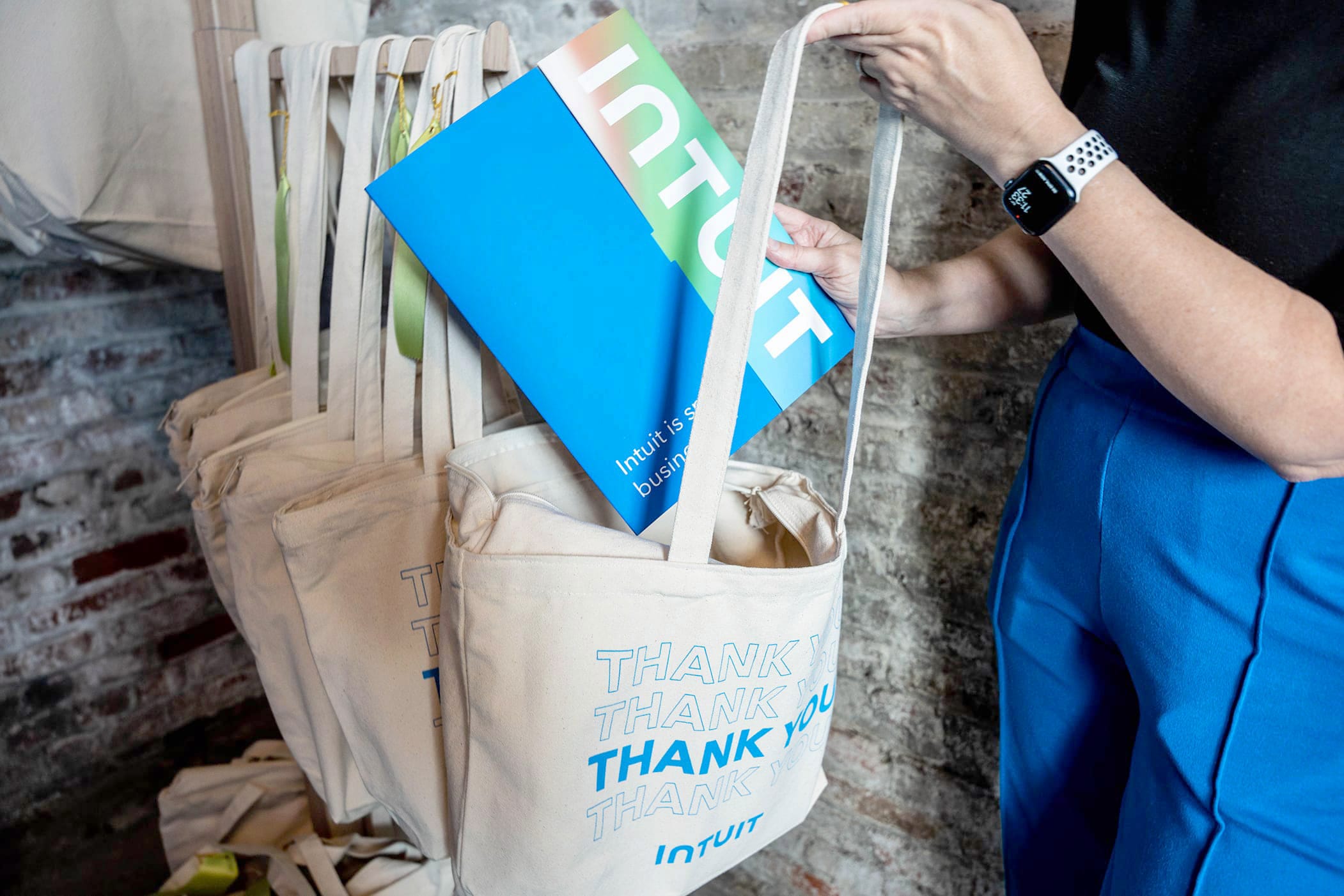 A person places an Intuit brochure into a beige tote bag marked with multiple "Thank You" messages in blue text. The tote bags are hanging in a row. The person is wearing a black top, blue pants, and a white smartwatch. A brick wall is in the background.