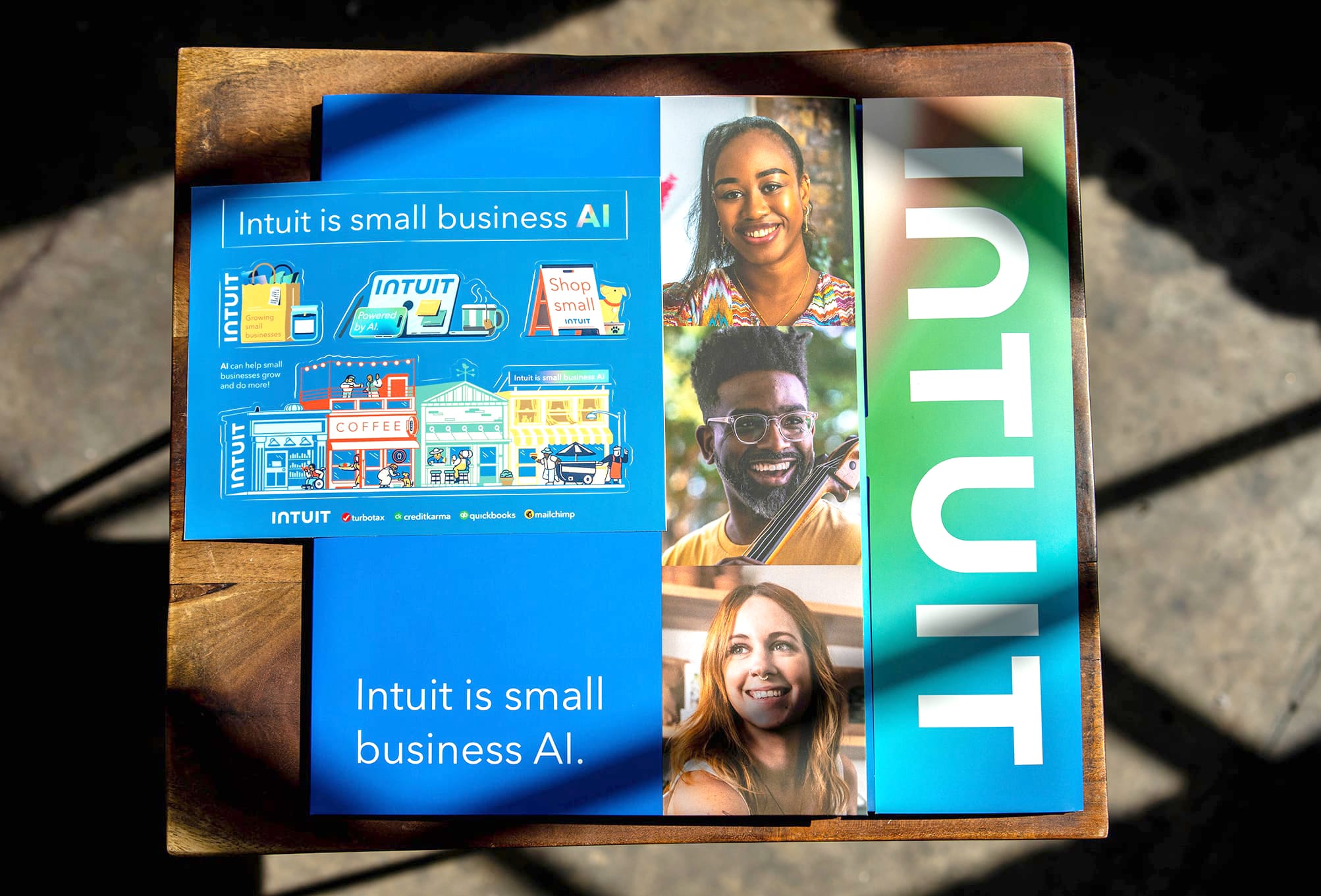 A blue Intuit brochure titled "Intuit is small business AI" lies on a wooden surface. It features images of a diverse group of smiling people and illustrations of small businesses, including a coffee shop. Various hashtags and social media icons are also visible.