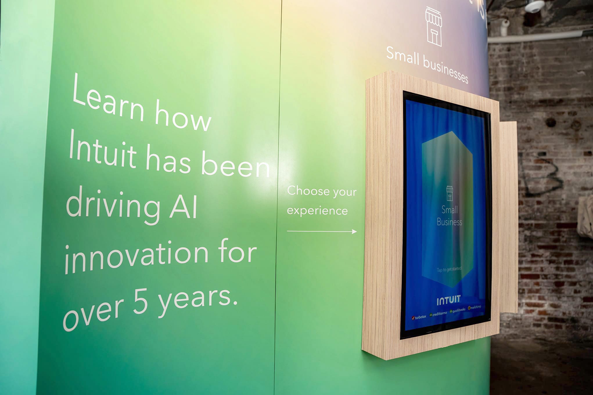 A green and yellow gradient wall displays the text, "Learn how Intuit has been driving AI innovation for over 5 years." To the right, there is a touchscreen framed in wood with a caption that reads, "Choose your experience," and an arrow pointing to the screen.