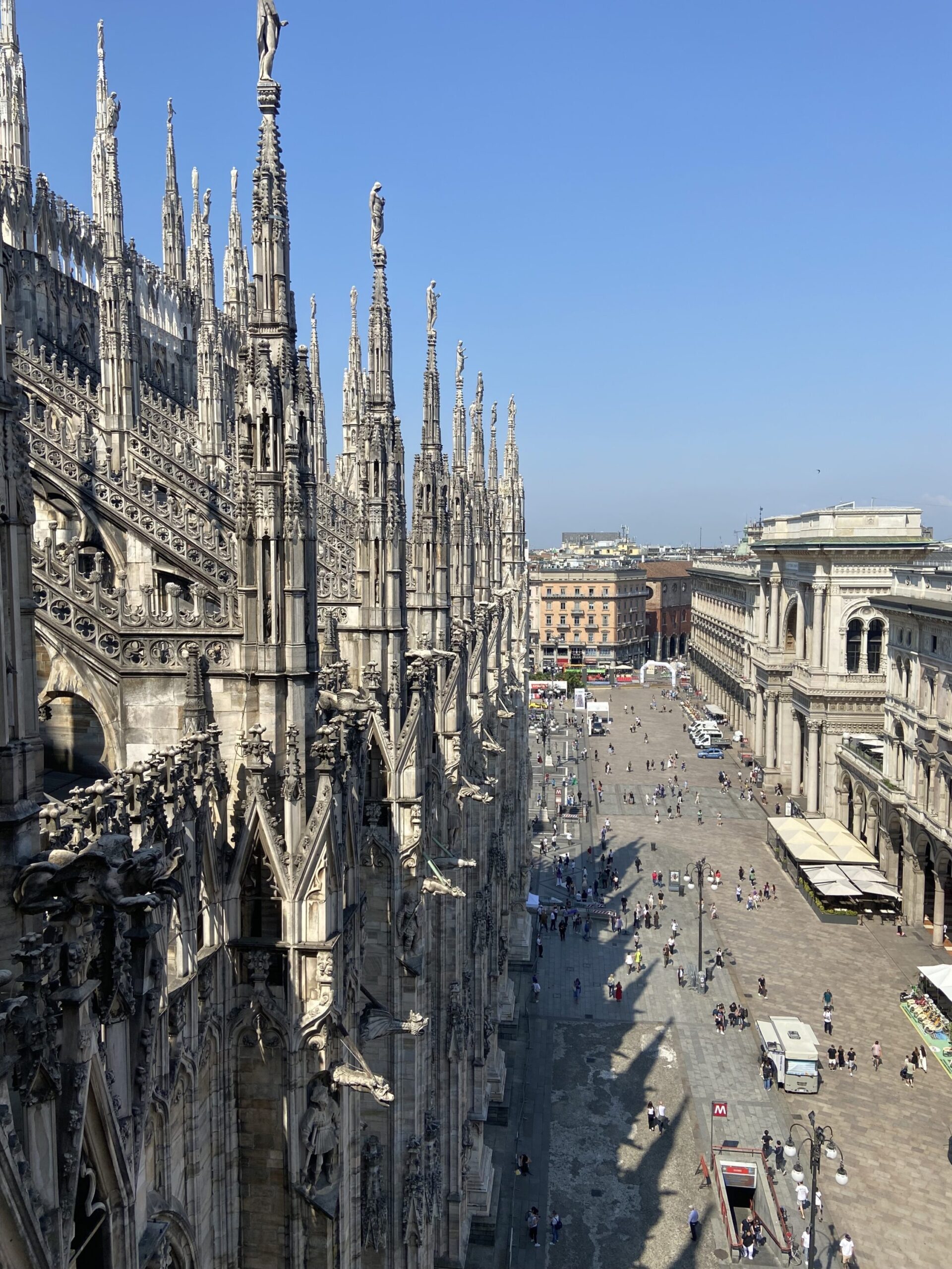 High-angle view of a large square in front of a cathedral, featuring intricate Gothic architecture with multiple spires. Many people are visible in the square, along with some outdoor seating, small structures, and vehicles. The sky is clear and blue.