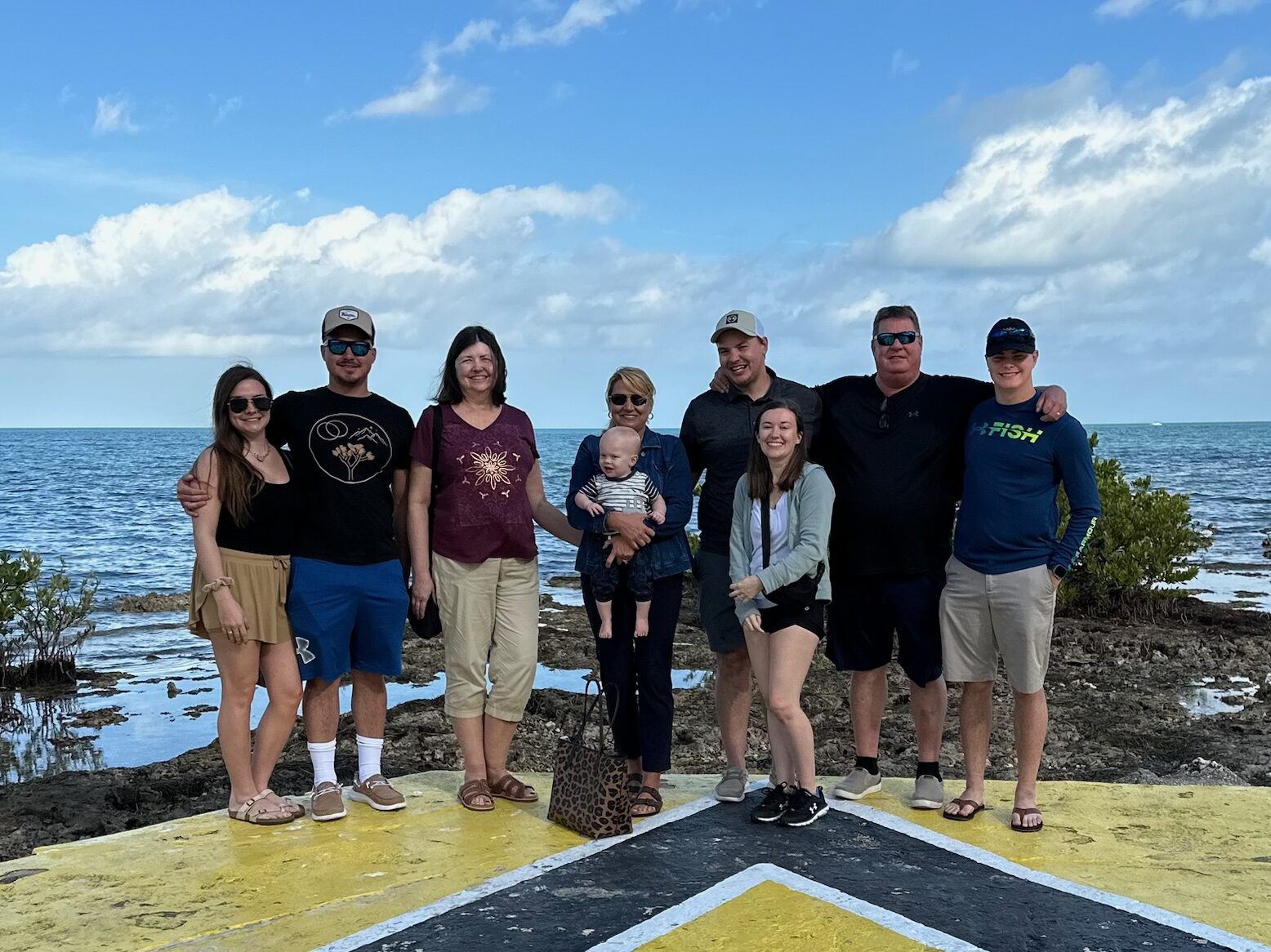 A group of nine people, including a baby, stand together on a yellow and black arrow painted on a concrete surface. They are in front of the ocean under a cloudy sky. Shrubbery and rocks can be seen along the shoreline. Everyone is smiling at the camera.