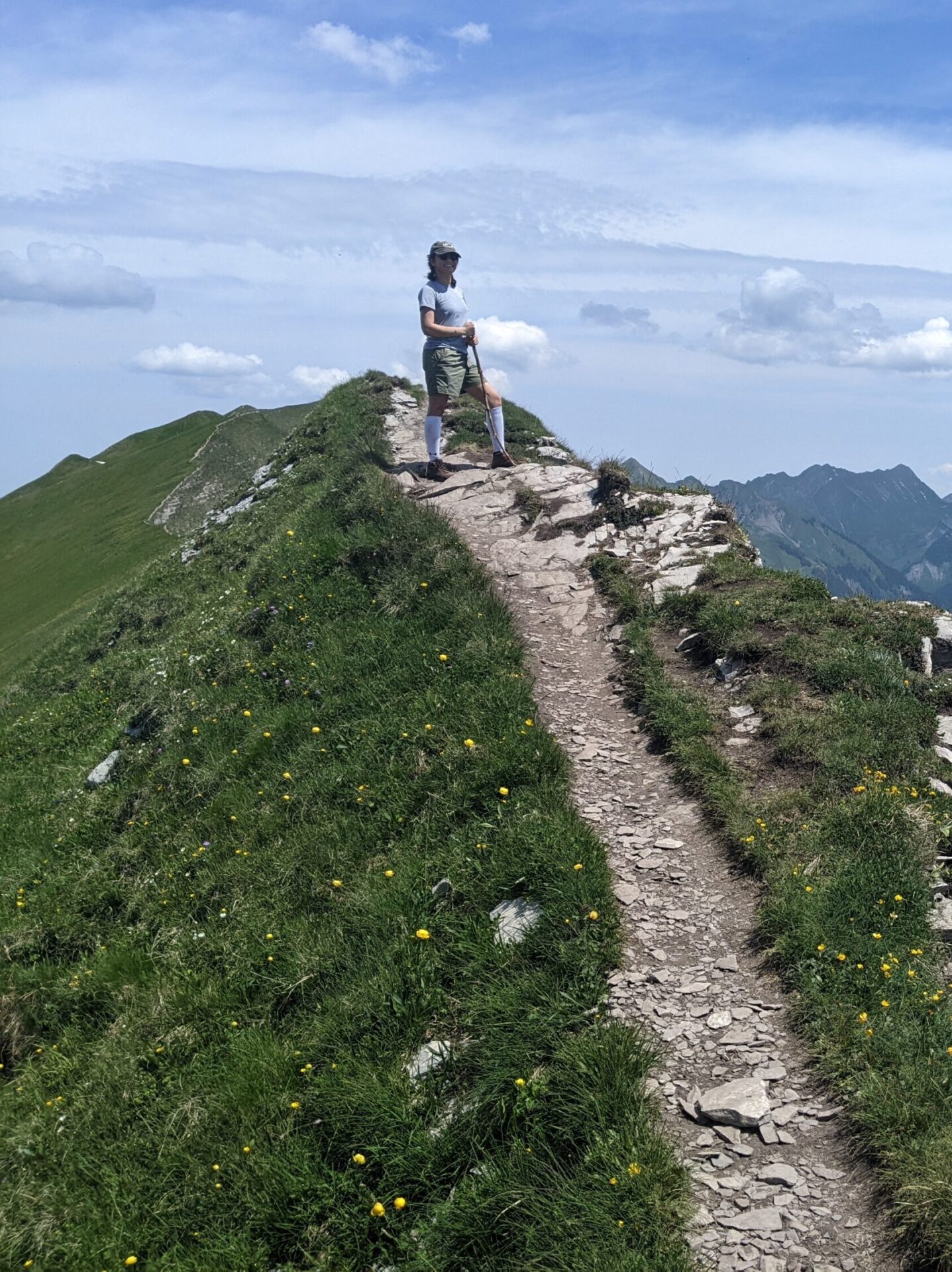 A person stands on a narrow, rocky mountain path with lush green grass on either side. In the background, there are expansive views of mountain ranges and a large, blue lake under a partly cloudy sky. The scene is serene and picturesque.