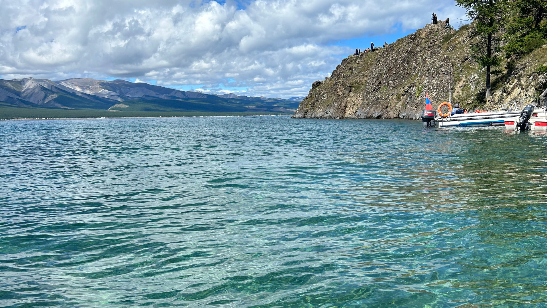 A serene lake with clear blue waters surrounded by mountains under a partly cloudy sky. A rocky hill on the right has people standing on top. Two motorboats are docked near the hill, with a lifebuoy visible on one boat. Pine trees are seen on the right.