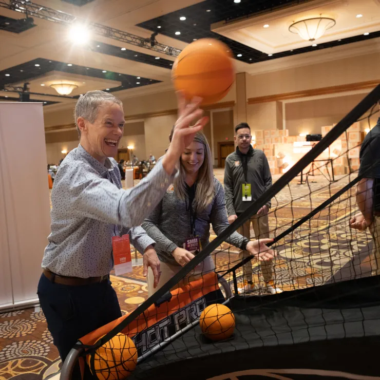 A person wearing a light blue shirt is playing an arcade basketball game in an indoor event setting. They are smiling as they shoot a basketball. Three other people are watching and smiling in the background. The floor is carpeted with a patterned design.