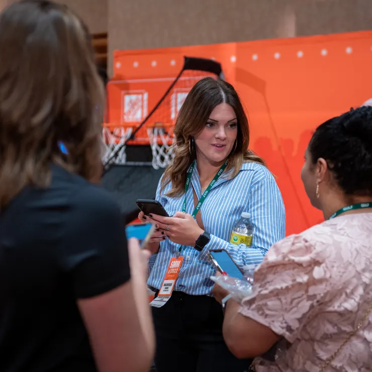 A woman in a blue striped shirt holds a phone and a water bottle while speaking to another woman in a pink blouse. They are at an event, and a basketball hoop with an orange backboard is visible in the background. Other attendees are partially visible in the foreground.