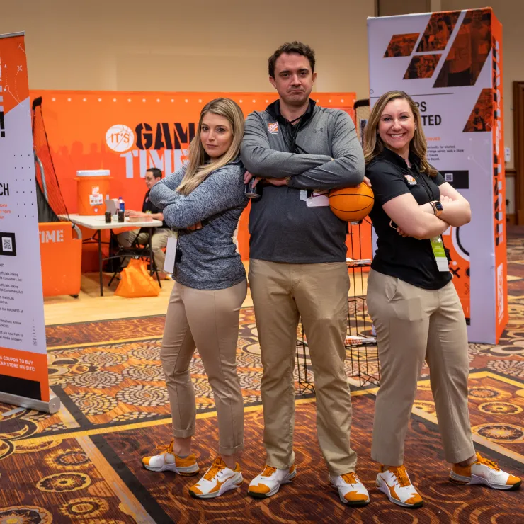 Three people strike confident poses with arms crossed, standing in front of a booth banner that reads "Game Time." They are indoors, on a patterned carpet. The two women are on either side of the man, all smiling and wearing casual attire, including sneakers.
