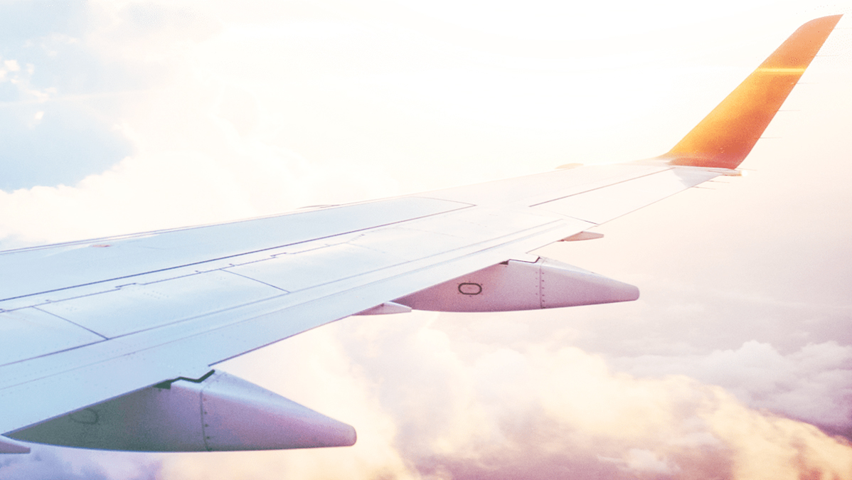 A view from an airplane window showing a wing with a gradient of orange light at the tip, against a backdrop of white clouds and a bright sky during sunrise or sunset. The sunlight casts a soft glow over the scene.