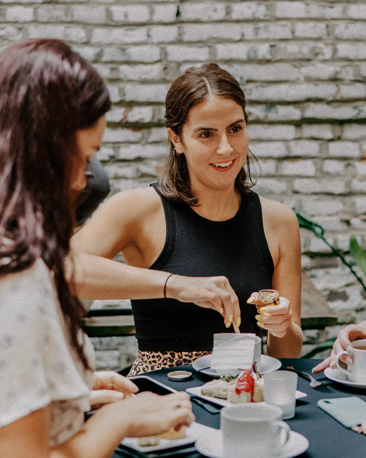 Three people sit at a table, enjoying a meal together. The woman in the center, wearing a black sleeveless top, is smiling and spreading a topping on her bread. The background features a brick wall, creating an outdoor or patio atmosphere.