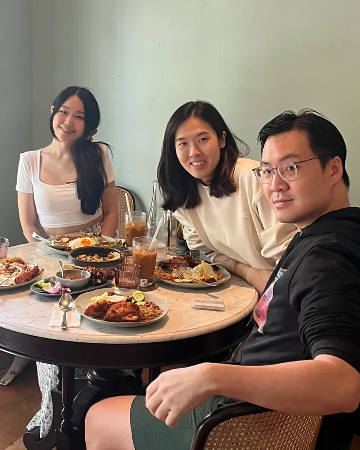 Three people sit around a round table in a cafe, smiling at the camera. The table is filled with various dishes and drinks, including iced coffee. The background shows a light green wall and wicker chairs. They appear to be enjoying a casual meal together.