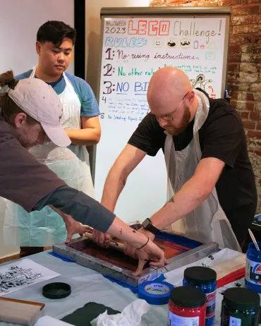Three individuals are engaged in a screen printing activity. One person is guiding another as they press a frame onto a print bed. Another person stands observing. Behind them, a whiteboard features a colorfully written "LEGO Challenge!" with three listed rules.