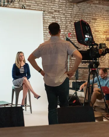 A woman sits on a stool in front of a white backdrop, being filmed by a cameraman. She wears a navy blazer and a black-and-white striped dress. Two men in the background adjust equipment and lighting in a brick-walled studio.