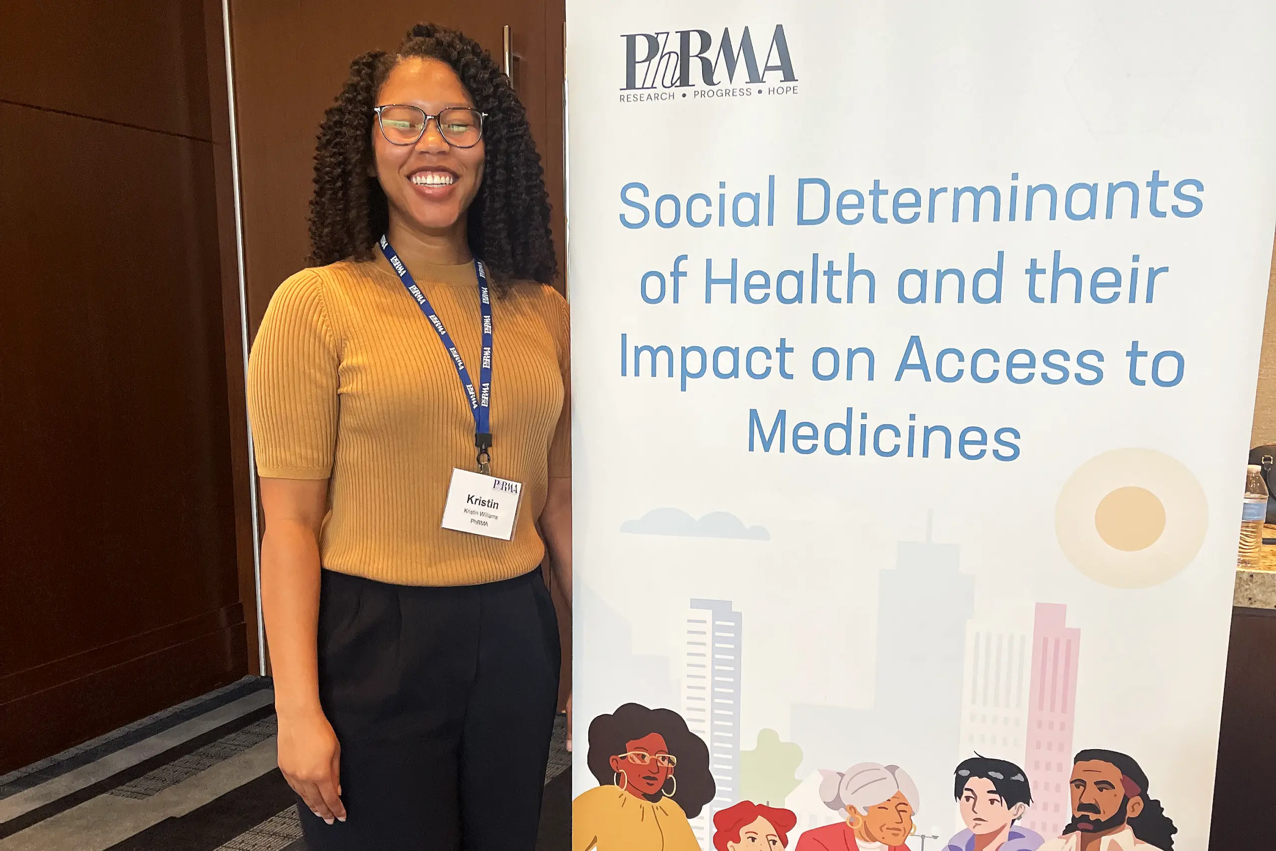 A person wearing a mustard-colored sweater and dark pants stands smiling beside a banner that reads "PhRMA - Research Progress Hope. Social Determinants of Health and their Impact on Access to Medicines." The banner has illustrations of diverse individuals.