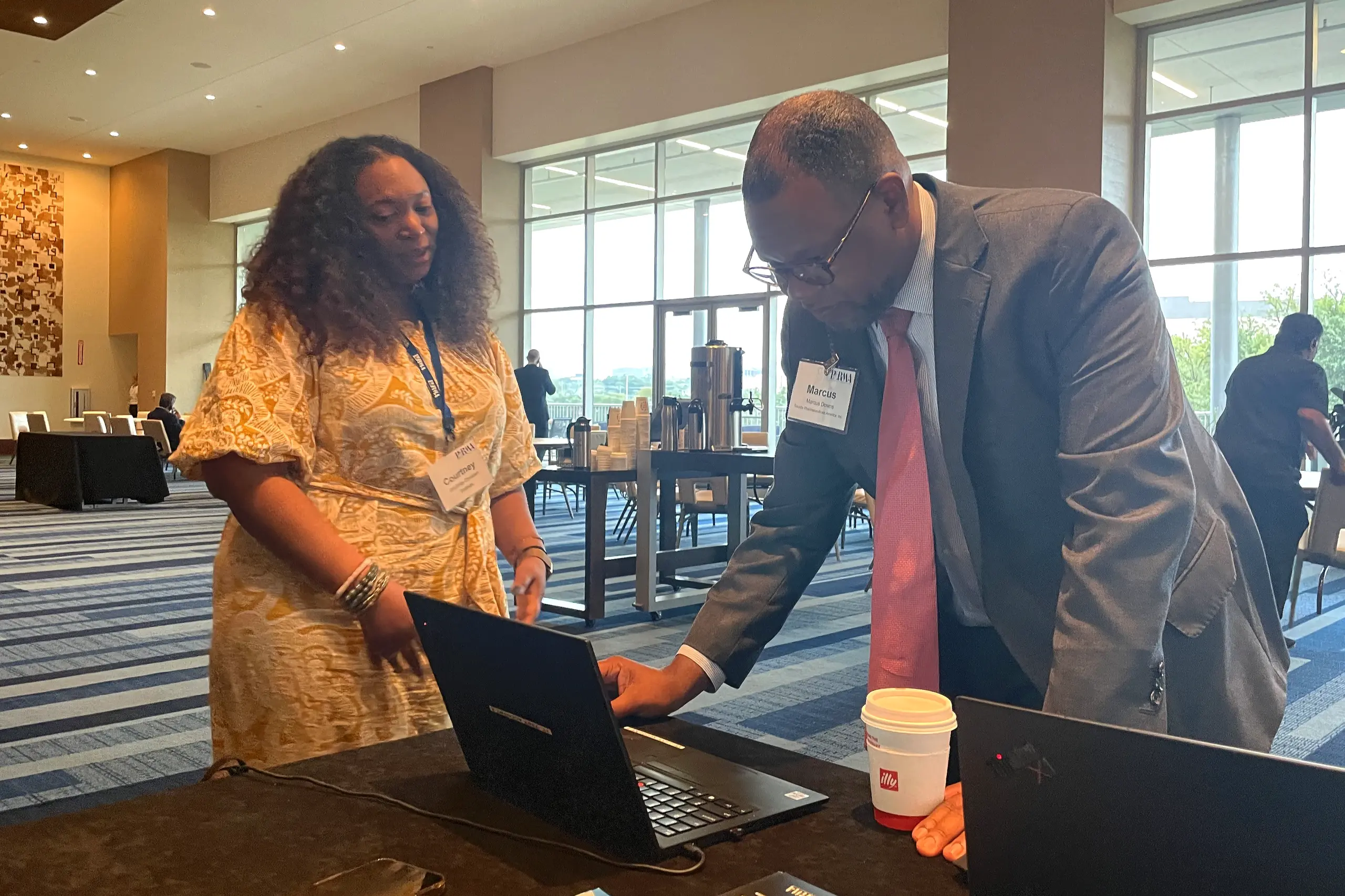 Two professionals are engaged in discussion at a conference booth. A woman in a yellow dress observes as a man in a suit and pink tie types on a laptop. Both wear name badges. The background features other attendees and large windows letting in natural light.