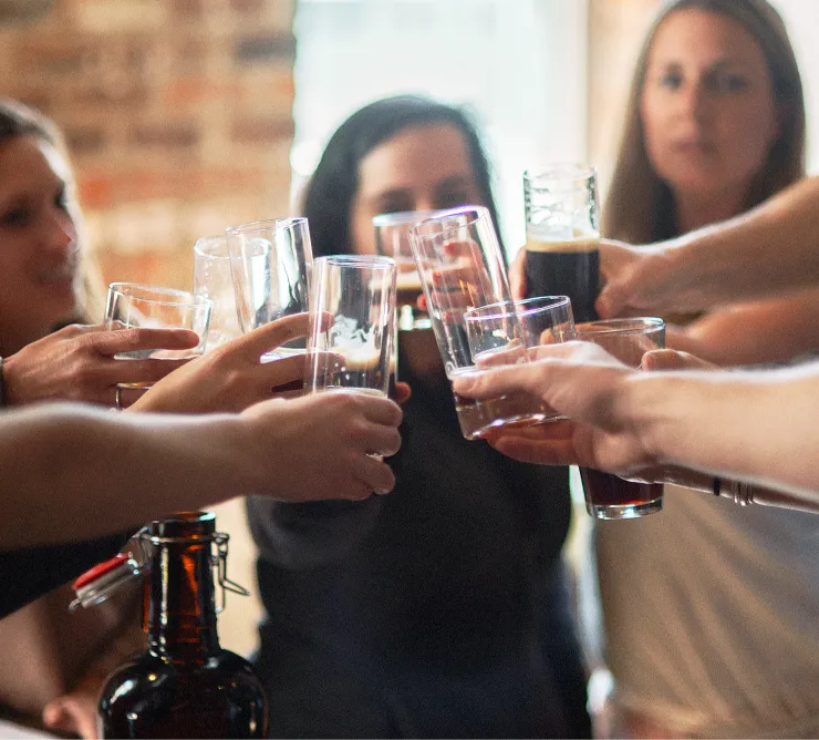 A group of people are clinking their glasses together in a toast. The scene appears to be indoors, with some blurred background elements like a brick wall. The glasses contain various beverages, and everyone seems to be celebrating or enjoying a social moment.