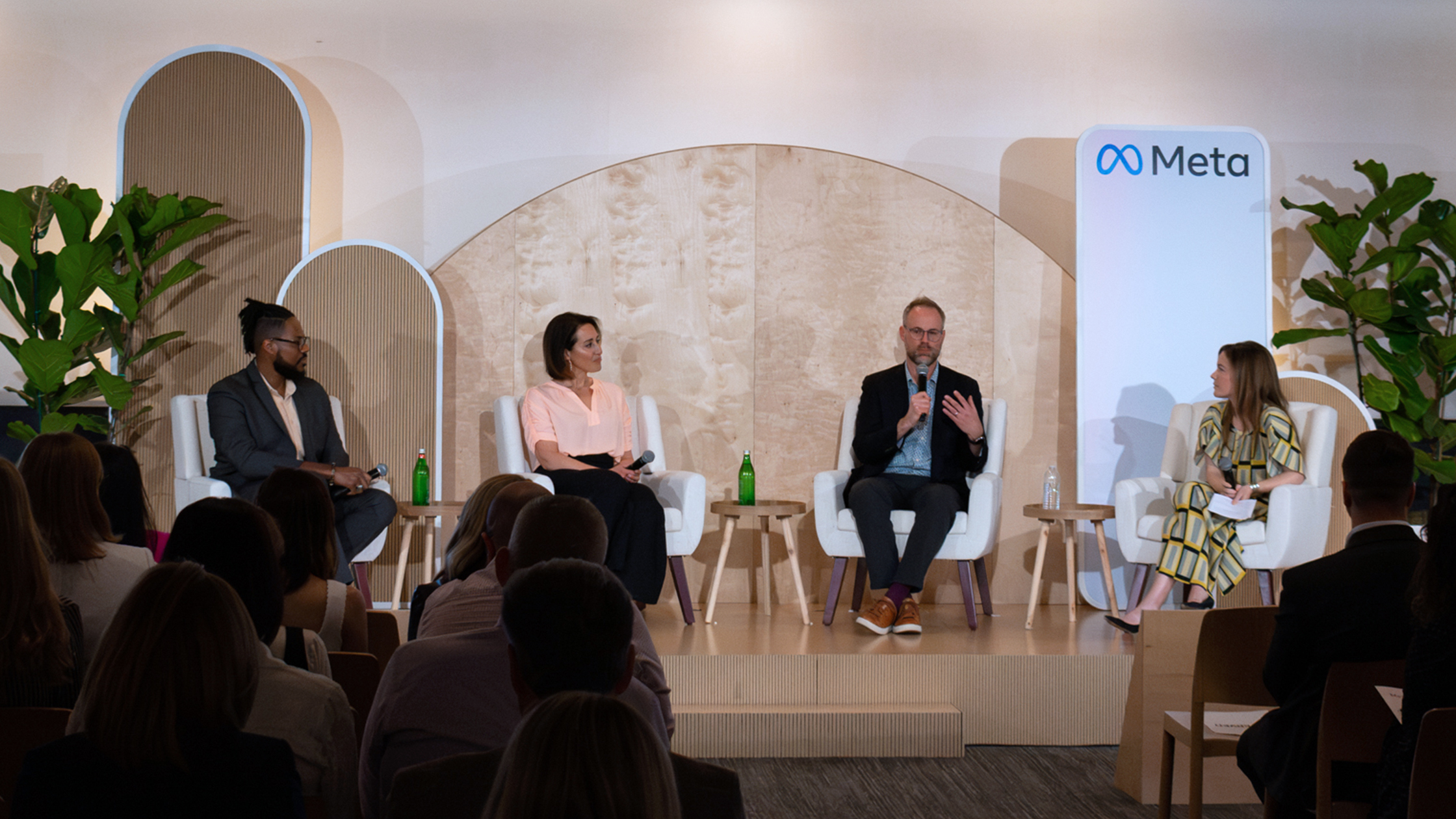 A panel of four people, two men and two women, are seated on a stage having a discussion. The man on the far right is speaking into a microphone. The stage features a Meta sign and potted plants. The audience is watching attentively.