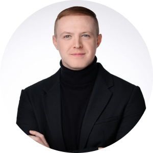 A person with short, light brown hair is wearing a black turtleneck and black blazer while standing against a plain white background. They are posing with their arms crossed and have a neutral expression on their face.