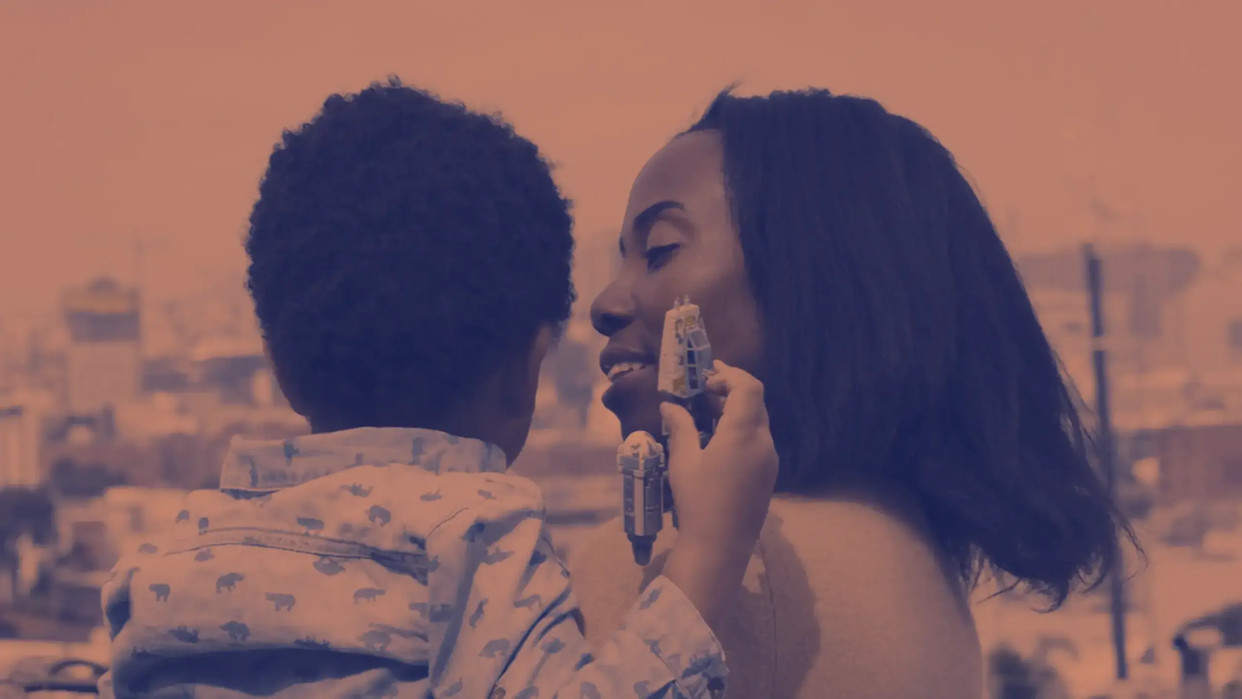 A young child with curly hair, wearing a patterned shirt, holds a toy car while looking up at a woman with shoulder-length hair. The woman smiles at the child while they stand outdoors with a blurry cityscape in the background. The image has a warm, muted color tone, reminiscent of creative storytelling in video production.