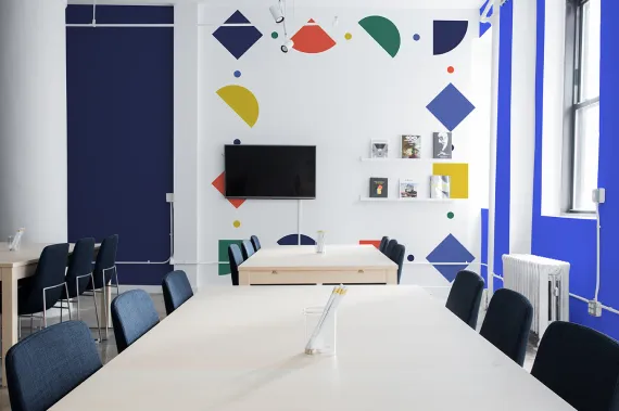 A brightly lit conference room featuring a long table with chairs around it. The white walls are decorated with colorful geometric shapes, and a flat screen TV is mounted on the wall. Books and magazines are displayed on modern white shelves to the right.