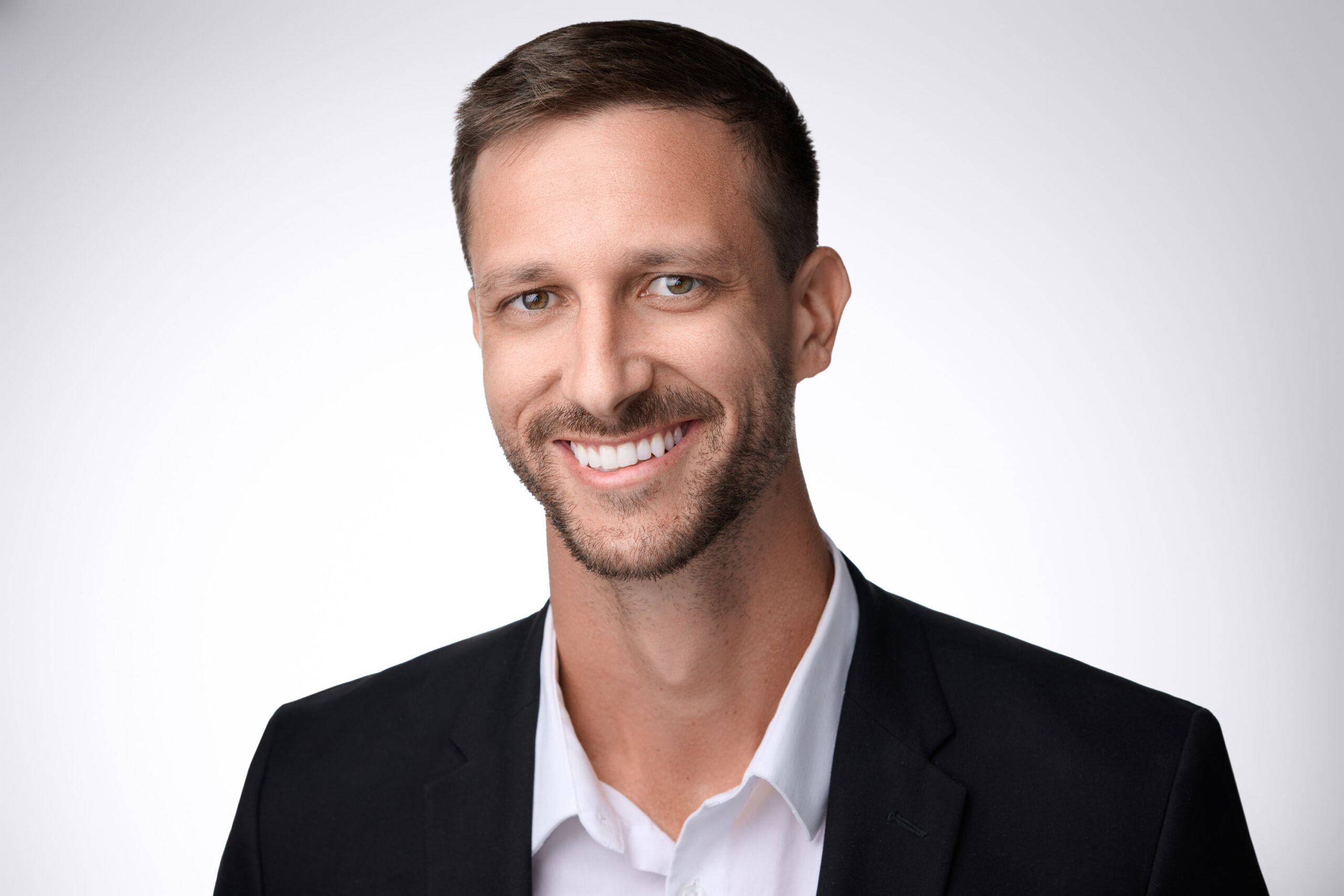 A man with short brown hair and a trimmed beard is smiling. He is wearing a black blazer over a white dress shirt. The background is plain white.