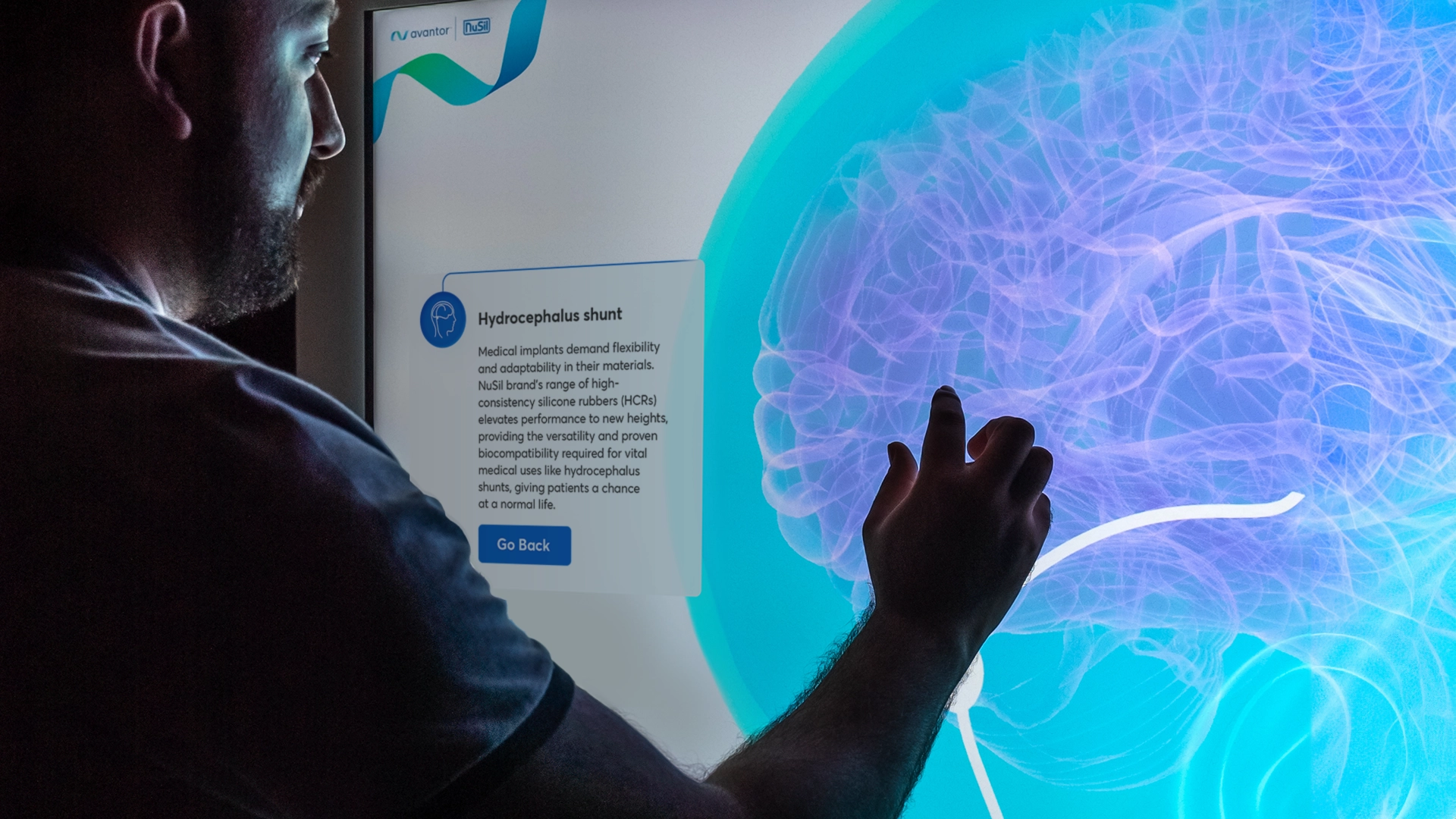 A person interacts with a touchscreen display illustrating a brain. The screen shows text about a "Hydrocephalus shunt," explaining medical requirements for materials to address conditions like hydrocephalus. The individual appears to be engaging with the information.