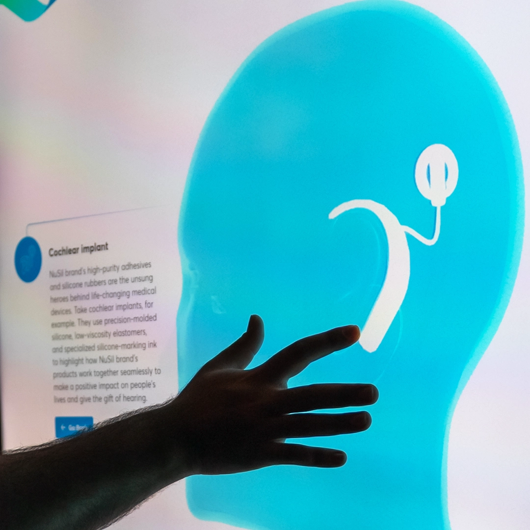 A silhouetted hand reaches towards a digital illustration of a human head showcasing a cochlear implant. The illustration is on a brightly lit screen, with text partially visible on the left side describing the cochlear implant technology.