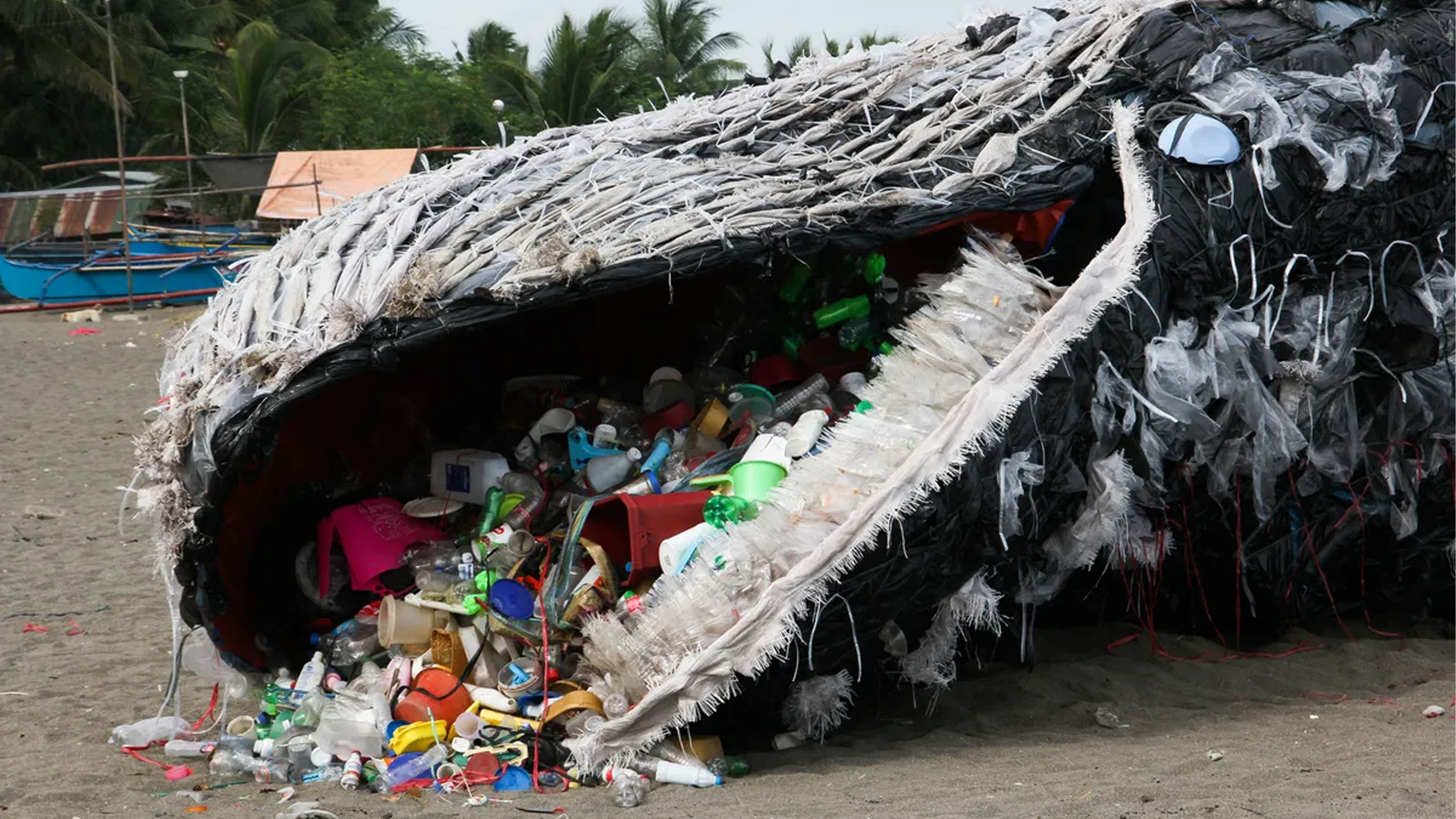 A large sculpture of a whale, intricately designed from plastic waste, sits on the beach. Its open mouth overflows with various plastic items, telling real stories from real people about environmental pollution. In the background, palm trees and parts of a boat add to the scene.