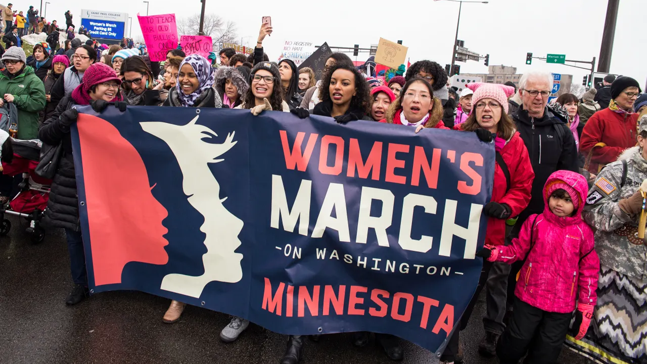 A diverse group of people march down the street, showcasing a design of unity with a large banner reading "Women's March on Washington Minnesota." Clad in winter clothing and holding colorful signs, they embody real stories from real people, creating an energetic and unified atmosphere.