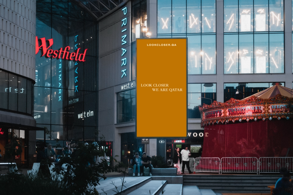 At dusk, a shopping center entrance glows with the vibrant branding of a brightly lit Primark store and gleaming Westfield signage. The scene is enhanced by a "Look Closer" ad and a festive red and gold carousel, capturing the essence of modern nation branding as people stroll past.