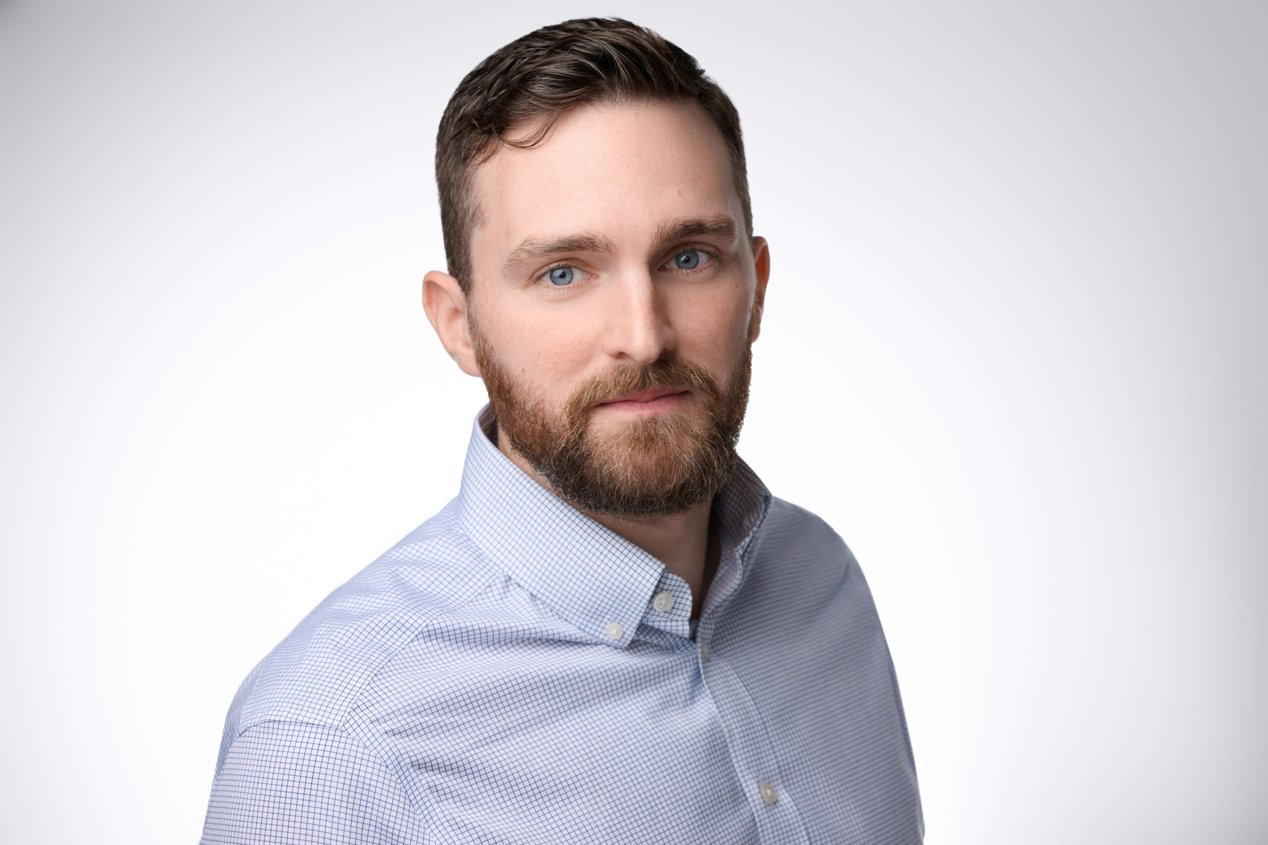Portrait of a man with short brown hair and a beard, wearing a light blue button-up shirt. He is facing slightly to the side, against a plain white background.
