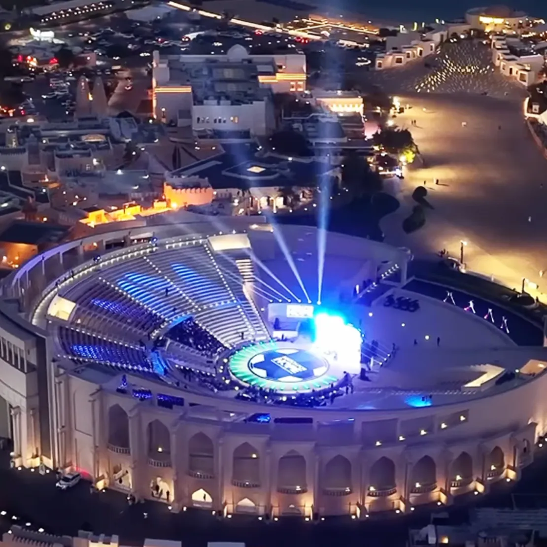 A large, illuminated outdoor amphitheater hosts an event at night, with blue and white lights shining on the stage and sparse audience seating—an ideal setting for public diplomacy events against the backdrop of a lively cityscape.