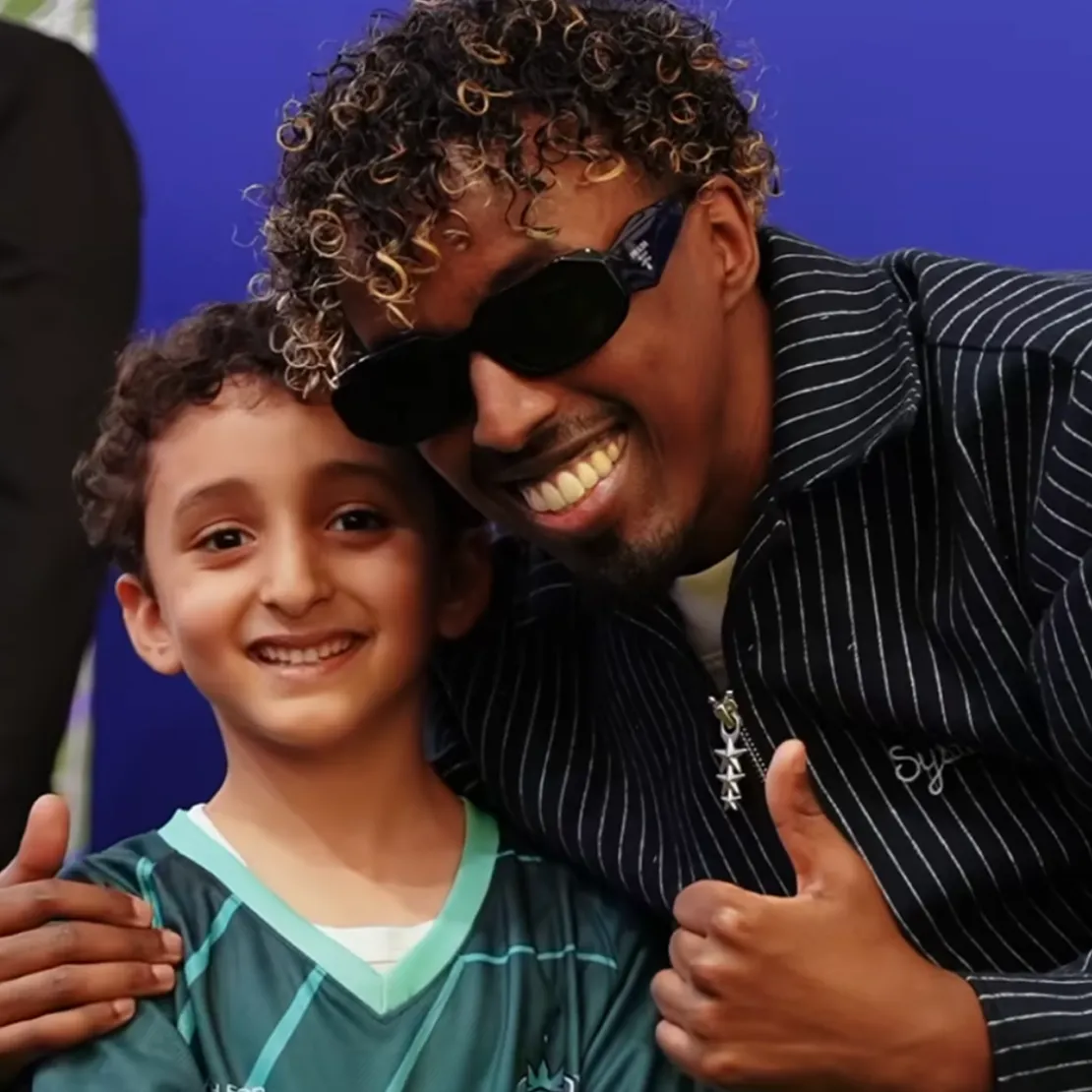 A smiling man in sunglasses and a striped jacket poses with his arm around a boy in a green sports jersey, both giving thumbs up in front of a blue background—celebrating the spirit of international relations.