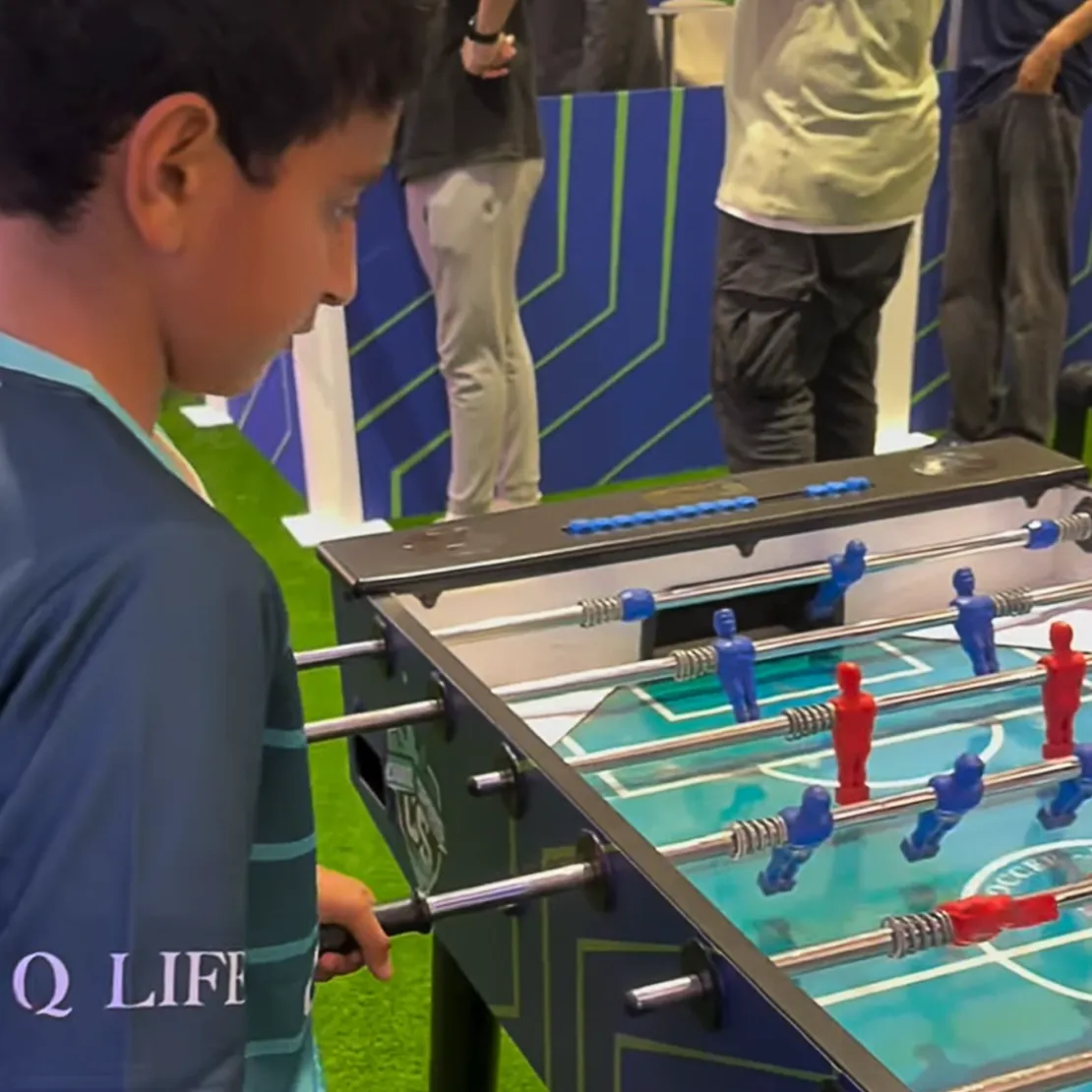 A boy plays table football, focusing on the game—a lively scene reminiscent of international relations, with blue and red players competing as others watch in the background.