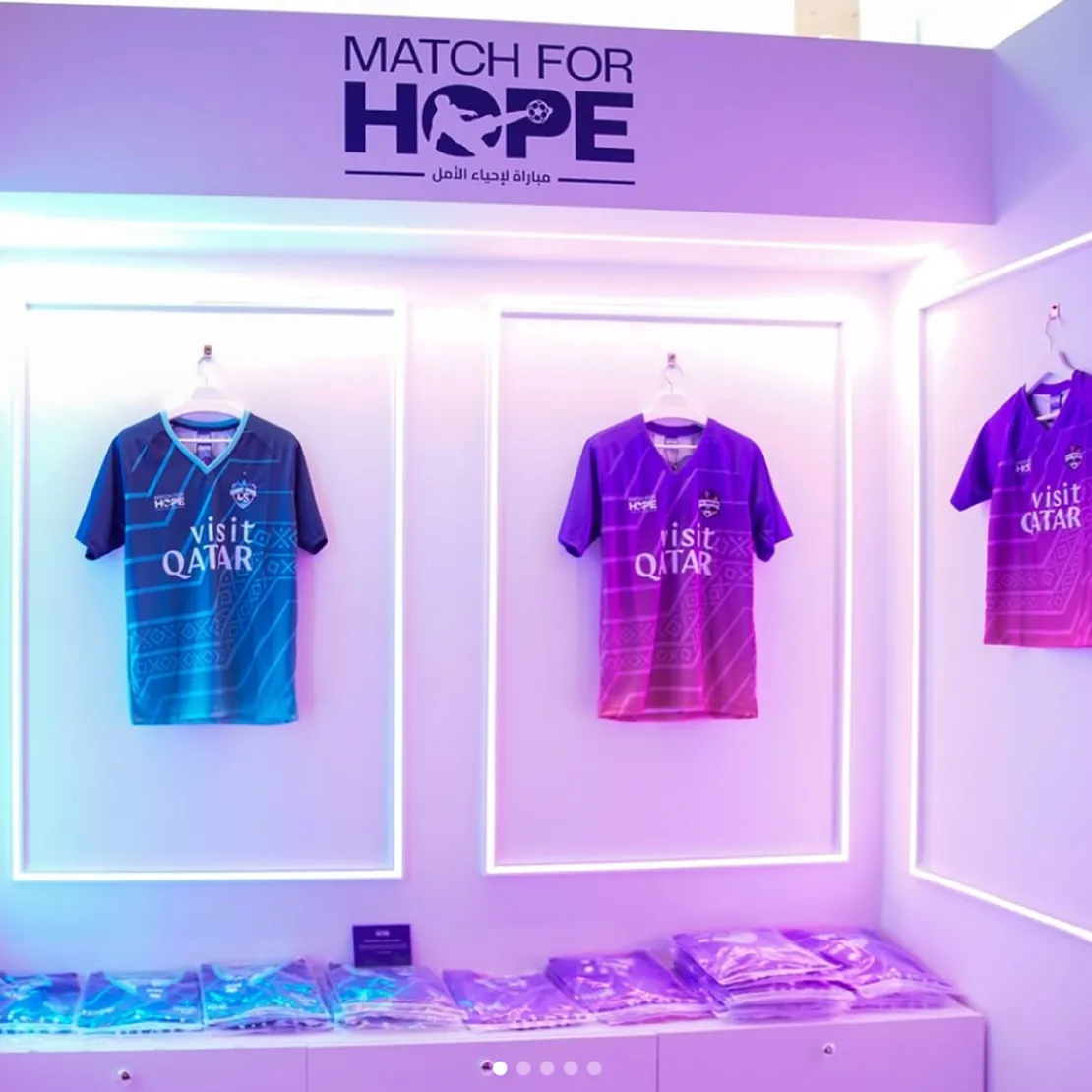 Three "Match for Hope" soccer jerseys—one blue, one pink, and one purple—are displayed on a white wall with LED lighting. Above them is a "Match for Hope" sign, highlighting this global campaign in nation branding. More jerseys are folded below.