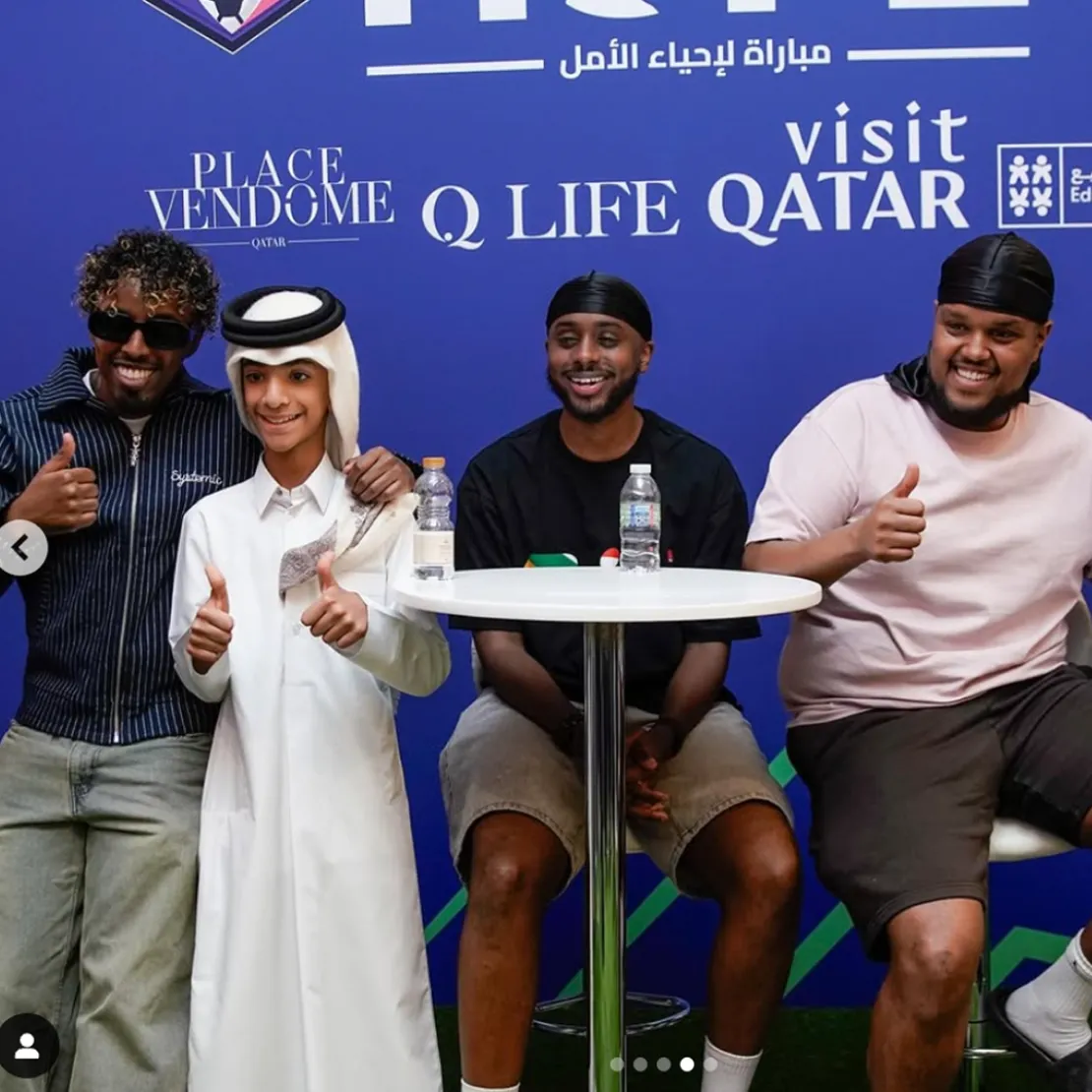 Four smiling men pose for a photo in front of a blue backdrop with “Q Life Qatar” and other logos, symbolizing nation branding. Two are seated at a round table, two stand, all giving thumbs up. One wears traditional Qatari attire.