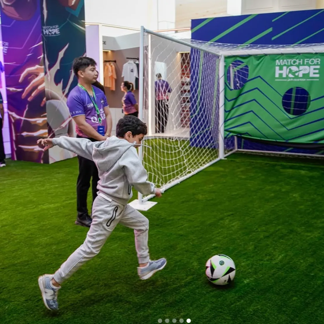 A young boy in a tracksuit kicks a soccer ball toward a goal at an indoor event area, part of the "Match for Hope" global campaign, while an adult stands nearby and watches.